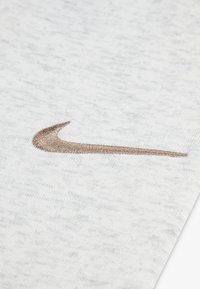 Lysegråt stof har et tekstureret, meleret mønster med en broderet, metalisk bronzefarvet Nike Swoosh-logo på overfladen.