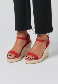 Red suede wedge sandals with a jute platform, featuring wide toe and ankle straps. The wedge height enhances stability and style.