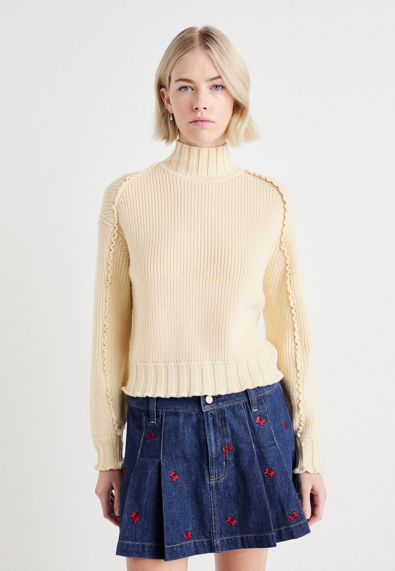 Pull à col roulé crème en côtes avec des détails texturés sur les manches, associé à une jupe en denim arborant une broderie de papillons rouges.