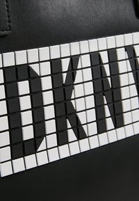 Černá taška s vzorovaným designem z malých černobílých dlaždic, které tvoří písmena a geometrické tvary na texturovaném povrchu.