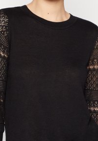 Schwarzer Pullover mit langen Spitzenärmeln, die ein Rautenmuster aufweisen. Weicher Stoff und klassisches Rundhalsausschnitt-Design. Keine weiteren Verzierungen.