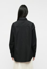 Camisa preta com botão, colarinho, mangas compridas e um corte descontraído. Textura suave, apresentando uma marcação discreta na pala das costas.