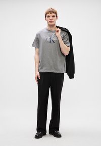 Γκρι κοντομάνικο μπλουζάκι με έντονο λογότυπο Calvin Klein, σε συνδυασμό με μαύρο παντελόνι ίσιας γραμμής και μαύρα παπούτσια, παρουσιάζοντας μια καθαρή, μοντέρνα γραμμή.