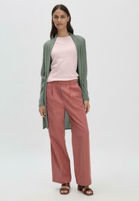 Lange, open groene cardigan over een lichtroze shirt, gecombineerd met hoogtaille, wijde roestkleurige broeken en bruine sandalen met hak.