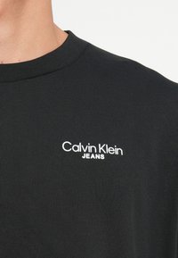 Crna pamučna majica s okruglim izrezom i rebrastom teksturom, s bijelim logotipom 'Calvin Klein JEANS' na lijevoj strani prsnog područja.