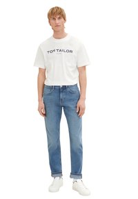 Tricou alb din bumbac cu text navy "TOM TAILOR", asociat cu blugi albaștri deschiși și teniși albi. Outfit simplu, casual cu blugi răsuciți.