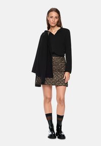 Schwarzes Langarmshirt, leopardgemusterter Minirock, schwarze Stiefeletten und durchsichtige Sportsocken. Das Model hält eine schwarze Jacke.