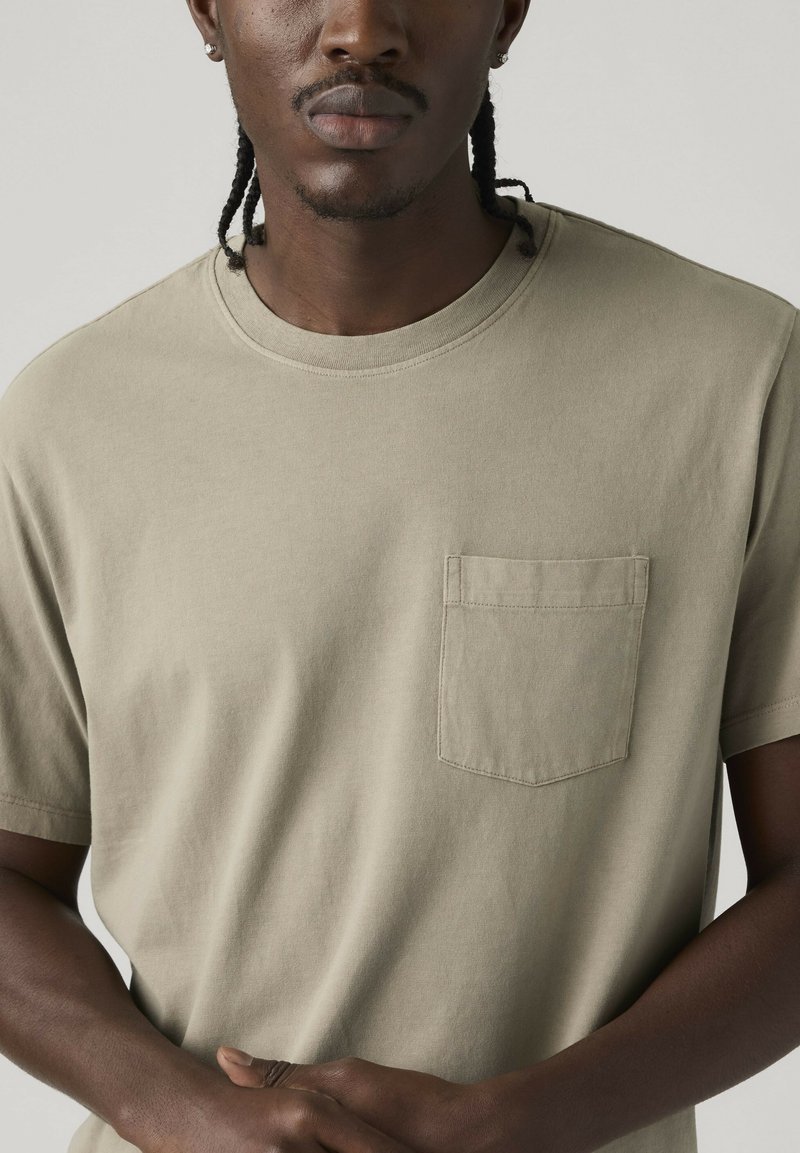 Homme aux cheveux tressés portant un t-shirt beige à manches courtes avec une poche poitrine, debout devant un fond clair uni.