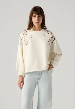Cream-colored sweatshirt with floral cut-out patterns on shoulders, round neckline, and relaxed fit, paired with light blue jeans.