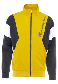 Chaqueta con cremallera amarilla y azul marino con acentos blancos. Cuenta con un cuello alto, bolsillos frontales y un logo en el pecho. Está hecha de un tejido ligero.