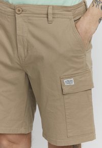 Khaki szorty cargo wykonane z bawełny o gładkiej teksturze, z zapięciem na guziki z przodu, bocznymi kieszeniami oraz naszywką na kieszeni cargo.