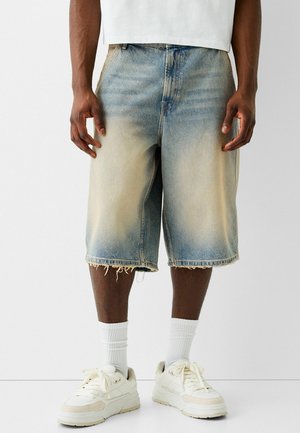 Homme portant un short en jean bleu clair délavé avec des ourlets effilochés, des chaussettes blanches montantes et des baskets blanches, debout devant un fond uni.
