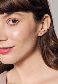 Srebrne kolczyki w kształcie sztyftów z małym czarnym kamieniem, osadzone w geometrycznym wzorze. Noszone w lewym uchu, prezentujące gładki, wypolerowany metal.