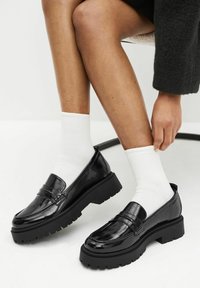 Zwarte patenten loafers met een dikke zwarte zool, gecombineerd met witte geribde sokken. De schoenen hebben een decoratieve naad over de wreef.