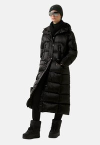 Schwarze Steppjacke mit langem Design, Kapuze, Reißverschlusstaschen und glänzender Textur. Kombiniert mit schwarzen Stiefeln und dunkler Kleidung.