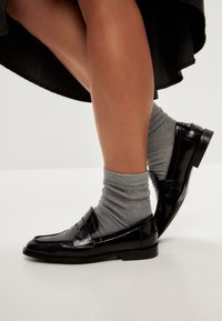 Svarta lackskinnsloafers med en klassisk design, i kombination med ljusgrå ankelsockor. Skon har en slät yta med minimal detaljering.