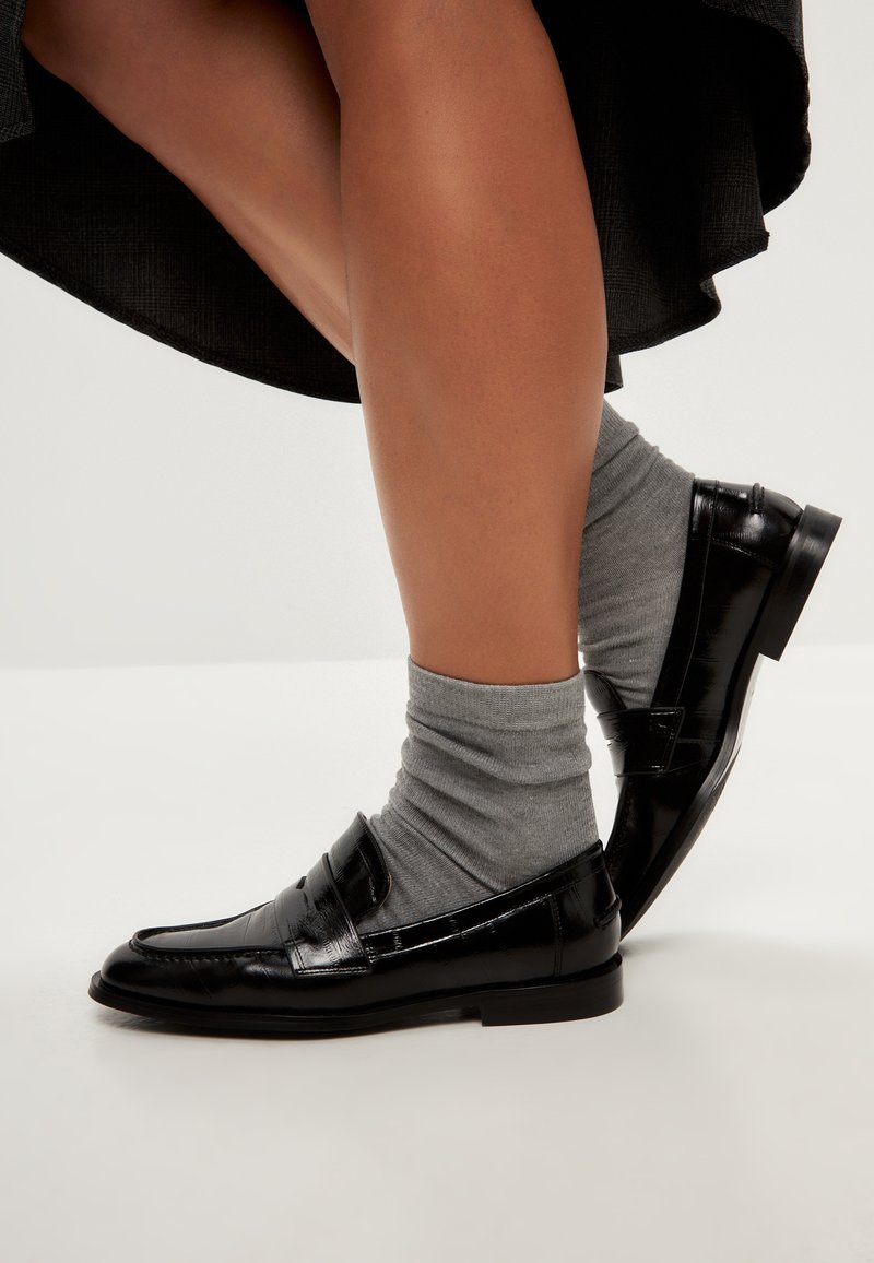 Svarta lackskinnsloafers med en klassisk design, i kombination med ljusgrå ankelsockor. Skon har en slät yta med minimal detaljering.