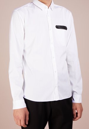Camisa blanca de botones con mangas largas, corte ajustado y un bolsillo negro con cremallera en el lado izquierdo del pecho. Textura de tela suave, cuello clásico.