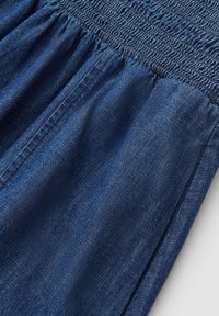 Donkerblauwe denim stof met een geplooide elastische tailleband en zichtbare naaddetails, getoond in een close-up tegen een lichte achtergrond.
