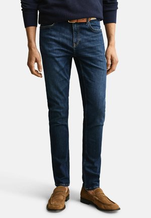 Mann trägt dunkelblaue Slim-Fit-Jeans, braunen Gürtel, marineblauen Pullover und braune Wildleder-Loafers, steht vor einem schlichten hellen Hintergrund.