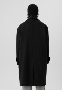 Persoon gezien van de achterkant, draagt een lange zwarte jas, een zwarte gebreide muts en lichtblauwe spijkerbroek tegen een effen grijze achtergrond.