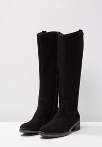 Une paire de bottes noires en daim montantes avec fermeture éclair sur le côté et un petit talon empilé, posées sur une surface blanche.