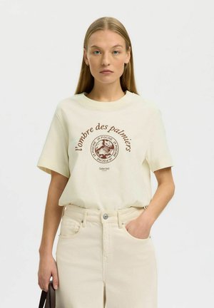 Nuori nainen suorilla vaaleanhiuksilla pukeutunut off-white-kuvioituun T-paitaan ja kermanvärisiin korkeavyötäröisiin farkkuihin, pitäen tummaa ruskeaa käsilaukkua toisessa kädessään.