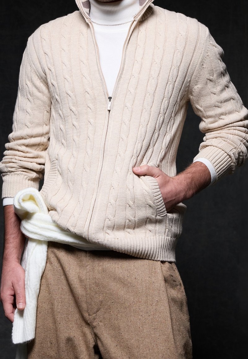 Homme portant un pull crème à torsades zippé sur un col roulé blanc, avec un pantalon beige et une écharpe blanche nouée autour de la taille.