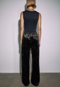 Osoba s krátkymi vlasmi, oblečená v bezrukávovom čiernom topi a širokých čiernych nohaviciach z mačkanej sametovej látky, stojí proti jednoduchej stene.