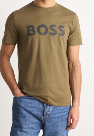 Hombre que lleva una camiseta verde oliva con "BOSS" en grandes letras negras, combinado con vaqueros azules, de pie con una mano en el bolsillo.