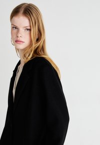 Svart oversized kåpe med en strukturert krage, mykt materiale og dype lommer, båret over en lys beige topp. Håret stylet i løse bølger.