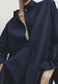 Chemise bleu marine boutonnée en tissu texturé, dotée d'un col pointu et d'une coupe décontractée, légèrement déboutonnée au niveau de la poitrine.