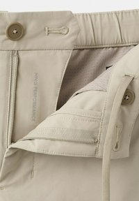 Pantalon beige avec fermeture à boutons, fermeture éclair et cordon de serrage réglable, montrant des coutures détaillées et une doublure intérieure avec de petites perforations.