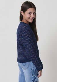 Maglione a maglia blu navy con motivo testurizzato e orlo a costine, abbinato a jeans azzurri con ricami floreali.