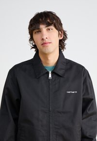 Veste noire en tissu durable, dotée d'une fermeture éclair, d'un col pointu et d'un petit logo blanc sur le côté gauche de la poitrine.