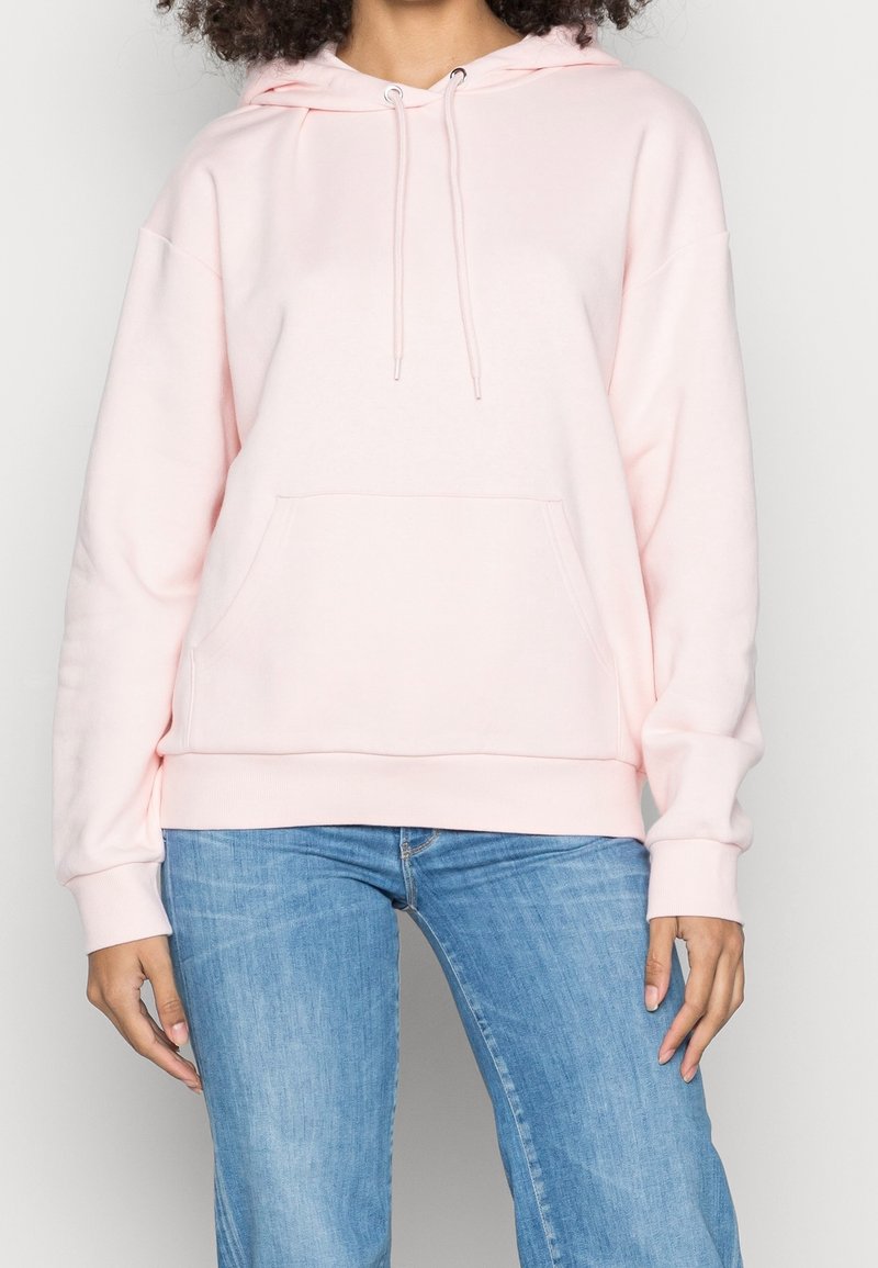 Donna con un hoodie rosa chiaro con tasca frontale e jeans azzurri, in piedi contro uno sfondo uniforme.