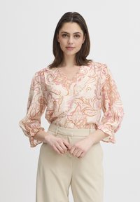 Blusa floreale paisley in tonalità di rosa chiaro e crema, con scollatura a V, maniche arricciate e tessuto leggero. Abbinata a pantaloni beige.