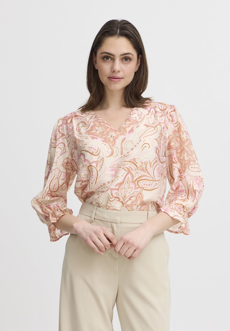 Blusa floreale paisley in tonalità di rosa chiaro e crema, con scollatura a V, maniche arricciate e tessuto leggero. Abbinata a pantaloni beige.