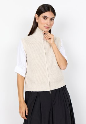 Frau mit langen dunklen Haaren, die eine beige gestrickte Weste mit Reißverschluss über einem weißen Hemd und einem schwarzen Rock trägt, posiert mit der Hand nahe am Kinn.