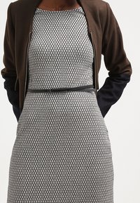 Veste zippée marron et marine portée sur une robe grise à motif avec une taille ajustée et une ceinture étroite. La robe présente une texture à motif losange.