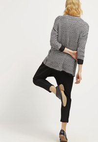 Blondýnka zvedající jednu nohu, oblečená v černém vzorovaném svetru, černých zkrácených kalhotách a černých lodičkách, otočená zády k jednobarevnému pozadí.