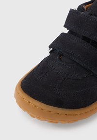 Μαύρο σουέντ αθλητικό παπούτσι με δύο λουριά βέλκρο και σόλα από καουτσούκ χρώματος καφέ. Στρογγυλή μύτη, υφή στην επάνω επιφάνεια και εμφανείς ραφές.