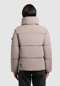 Lys beige pufferjakke med høj krave, der har horisontale quiltede sektioner, en glat overflade og et sort brandlogo på siden.