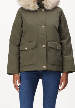 Olivenfarvet vinterjakke med store knapper, to forlommer med klap og hætte foret med pels, båret sammen med blå jeans.