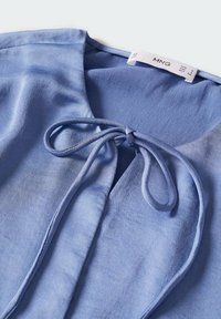 Blouse en tissu doux bleu avec un col rond et un détail de nœud noué au col avant, montrant une étiquette de vêtement à l'intérieur.