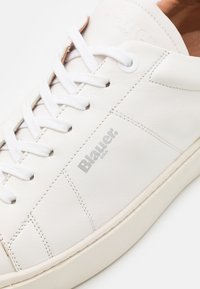 Sneaker in pelle bianca con lacci bianchi, caratterizzato da una superficie texturizzata e cuciture visibili. Marchiato "Blauer USA" sul lato esterno.