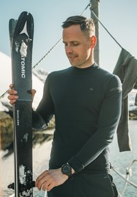 Uomo con camicia nera a maniche lunghe che aggiusta uno sci Atomic nero con macchie di neve, vicino a una riva rocciosa innevata e all'acqua sotto un cielo sereno.