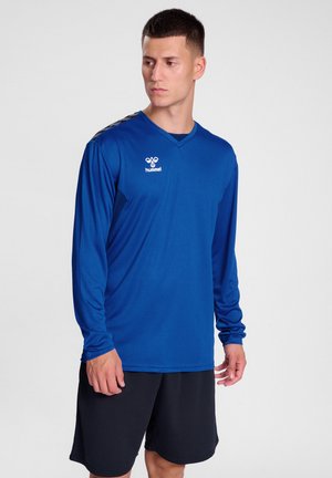 Langarm blauwe sportshirt met V-hals, voorzien van zwarte accenten op de schouders en een logo op de borst. Gecombineerd met zwarte shorts.