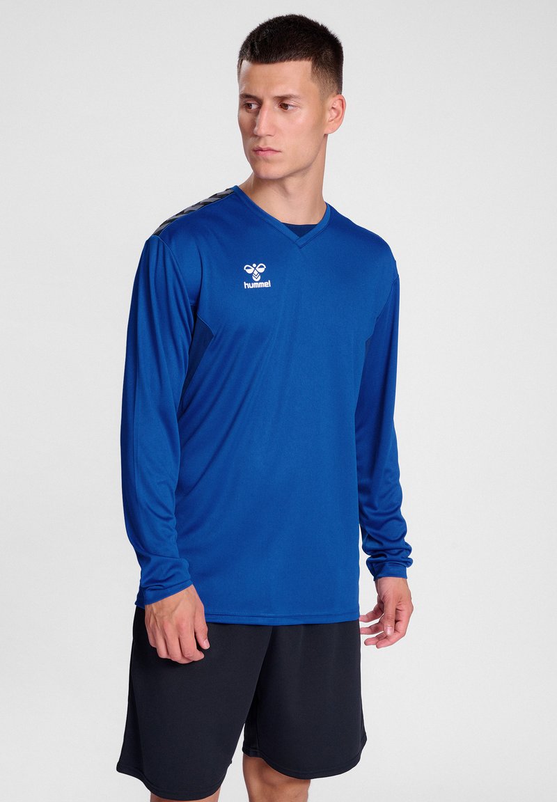 Langärmliges blaues Sportshirt mit V-Ausschnitt, mit schwarzen Akzenten an den Schultern und einem Logo auf der Brust. Kombiniert mit schwarzen Shorts.