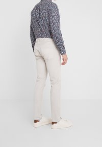 Jasnobeżowe spodnie o dopasowanym kroju, wykonane z miękkiego materiału, zestawione z ciemną, wzorzystą koszulą oraz białymi sneakersami z brązowymi akcentami.